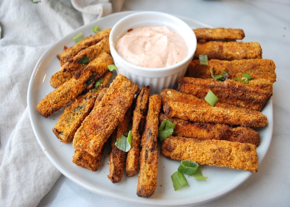 Plate of air fryer zucchini fries with a white pot of yogurt dipping sauce.